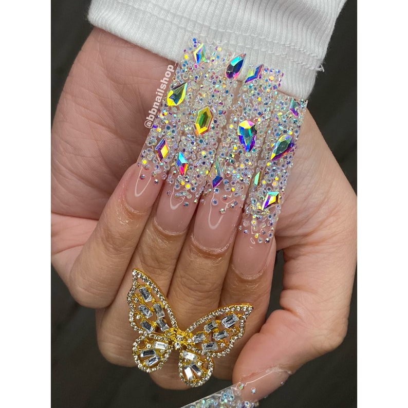 May include: Long, clear acrylic nails with a butterfly ring. The nails are decorated with iridescent rhinestones and small white beads.