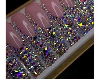 Peut inclure: Un ensemble de dix faux ongles roses avec des strass irisés disposés en cascade. Les ongles sont présentés dans une boîte blanche avec le texte "@bbnailshop_" en bas à gauche.
