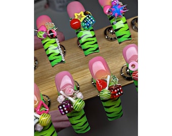 Puede incluir: Una colección de uñas postizas largas con un diseño de rayas de tigre rosa y verde. Cada uña presenta un elemento decorativo diferente, incluyendo dados en miniatura, estrellas y flores. El texto "bbnailshop" es visible en una uña.