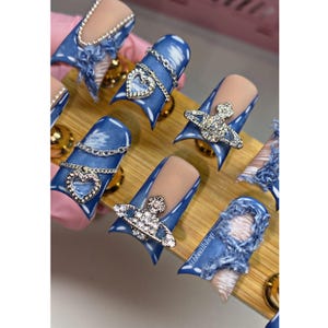 May include: A set of blue and white faux nail tips with silver charms. The charms include a heart and a small circle with rhinestones. The nails are displayed on a wooden surface.