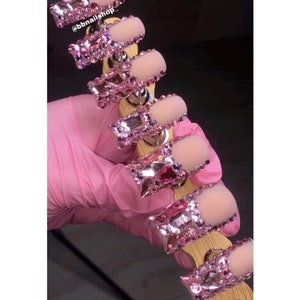May include: A set of pink press-on nails with a silver base and pink rhinestones. The nails are arranged on a wooden stick with a silver base. The text "@bbnailshop_" is visible in the image.