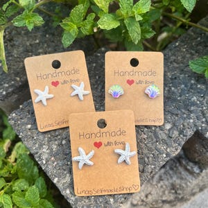 Könnte beinhalten: Drei Paar Ohrstecker auf braunen Pappkarten mit der Aufschrift Handmade with love und LinasSefrmadeShop. Zwei Paare zeigen weiße, texturierte Seesterne und ein Paar irisierende Muschelformen mit einer perlmuttartigen Oberfläche.