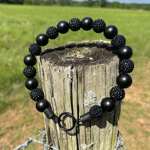 May include: Black beaded dog collar featuring a mix of smooth and textured beads. The collar has a metal ring for leash attachment and is displayed outdoors against a natural backdrop of a wooden post and green field.