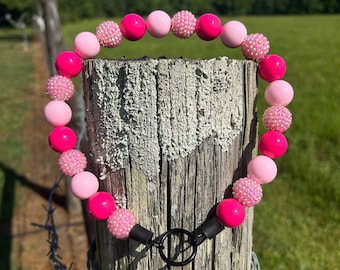 May include: A pink beaded collar with a black clasp, featuring various shades of pink beads. Some beads have a textured, sparkly surface. The collar is displayed on a weathered wooden post with barbed wire.