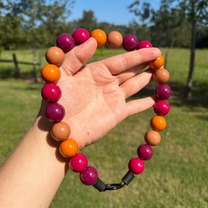 May include: A colourful necklace made of round beads in shades of orange, pink, and burgundy. The necklace is strung on a black cord with a metal clasp.
