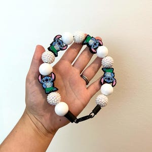 May include: A beaded pacifier clip featuring Stitch character charms. The clip has alternating white and textured white beads, with black connectors and a black clip. The Stitch charms are blue, green, and pink.