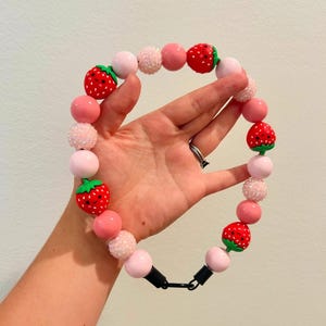 May include: A pink and red beaded necklace featuring strawberry and round bead accents. The necklace has a black clasp and is held in a hand. The beads are in shades of pink and white.