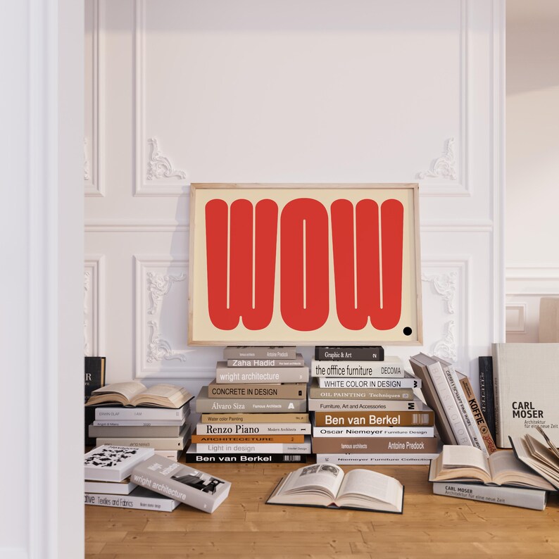 May include: A framed art print with the word "WOW" in bold red letters on a cream background. The artwork is displayed above a collection of books on a wooden surface, creating a stylish interior design scene.