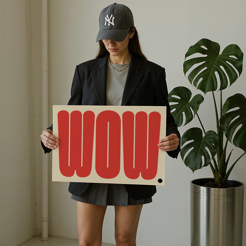 May include: A rectangular sign with the word "WOW" in bold, red letters. The sign is held by a person wearing a dark gray baseball cap with a white logo, a black blazer, and a gray t-shirt. A large potted plant is in the background.