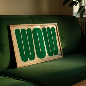 May include: A framed art print featuring the word "WOW" in bold, green, rounded letters on a cream background. The artwork is displayed on a green sofa, with a small blue dot in the lower right corner.