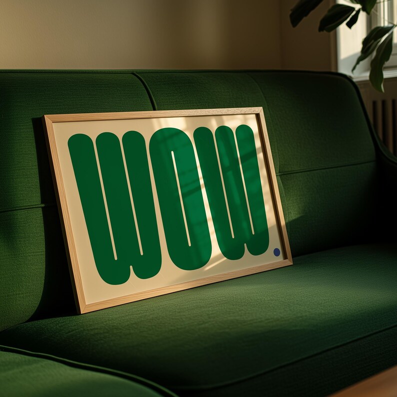 May include: A framed art print featuring the word "WOW" in bold, green, rounded letters on a cream background. The artwork is displayed on a green sofa, with a small blue dot in the lower right corner.