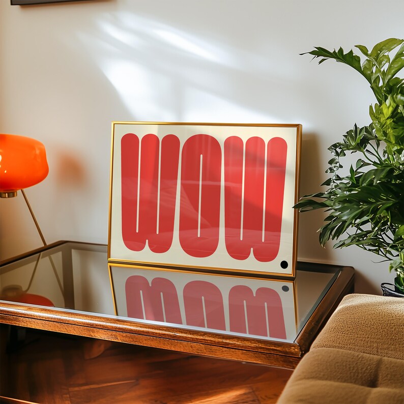 May include: A framed art print with the word "WOW" in bold, red letters on a cream background. The artwork is displayed on a mirrored surface, reflecting the design. The frame is gold-colored.