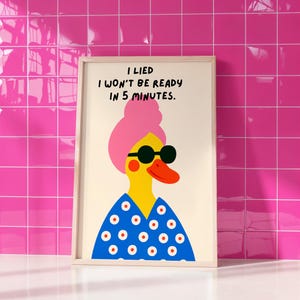 以下が含まれることがあります： サングラスとピンクのヘアバンドをつけた漫画のアヒルの額装アートプリント。アヒルは黄色い顔と赤いクチバシをしており、白い水玉模様の青いトップスを着ています。プリントのテキストは「嘘をついた、5分で準備はできません」と書かれています。