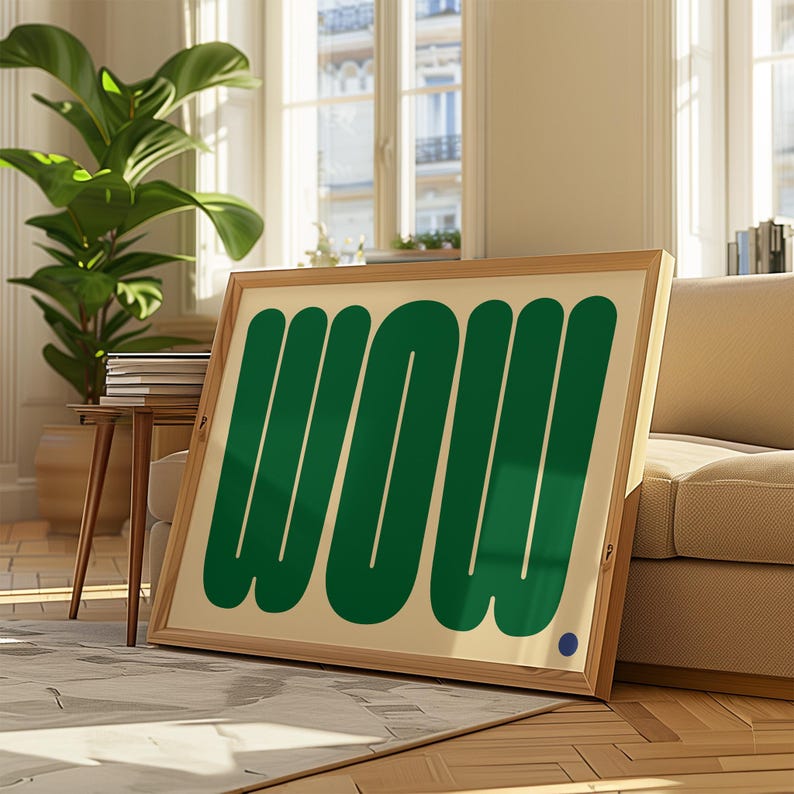 May include: A framed art print with the word "WOW" in bold, green letters on a cream background. The wooden frame is light brown. A small blue dot is in the lower right corner. The artwork is leaning against a beige sofa.