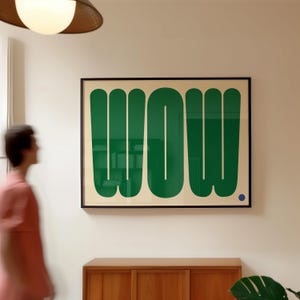 May include: A framed art print with the word "WOW" in large, green, stylized letters. The artwork is set against a cream background and framed in black. A small blue dot is in the lower right corner.