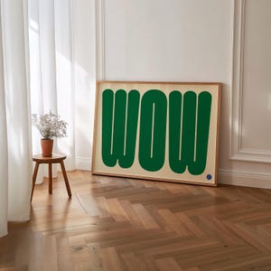 May include: A framed art print featuring the word "WOW" in large, green, rounded letters against a cream background. The artwork is in a light wood frame and leans against a white wall. A small blue dot is in the lower right corner.