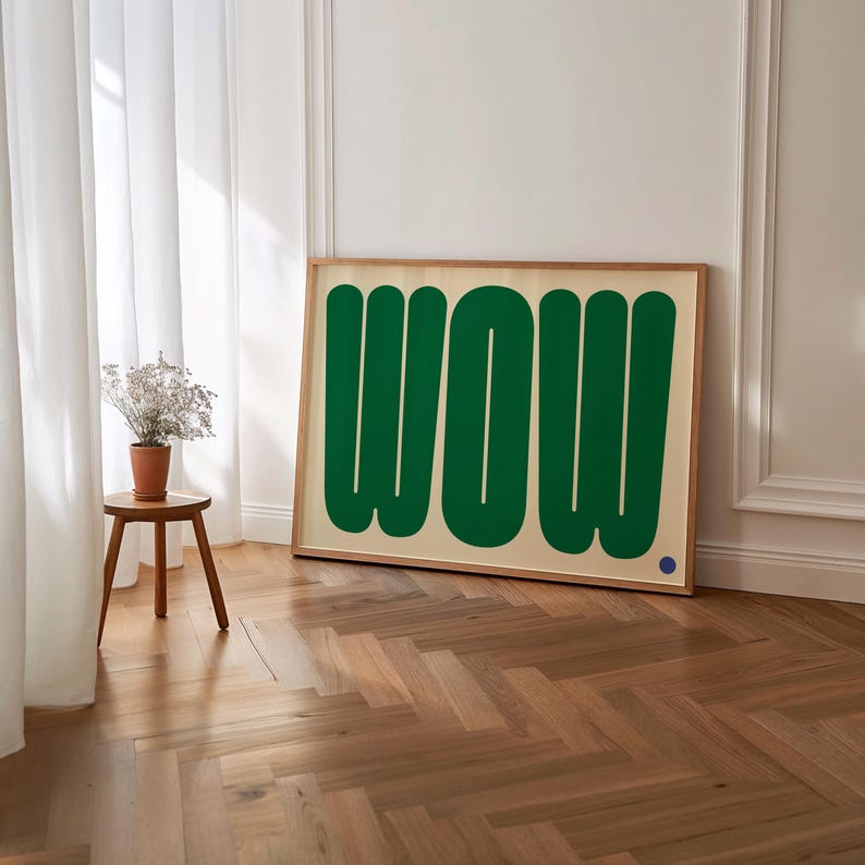 May include: A framed art print featuring the word "WOW" in large, green, rounded letters against a cream background. The artwork is in a light wood frame and leans against a white wall. A small blue dot is in the lower right corner.