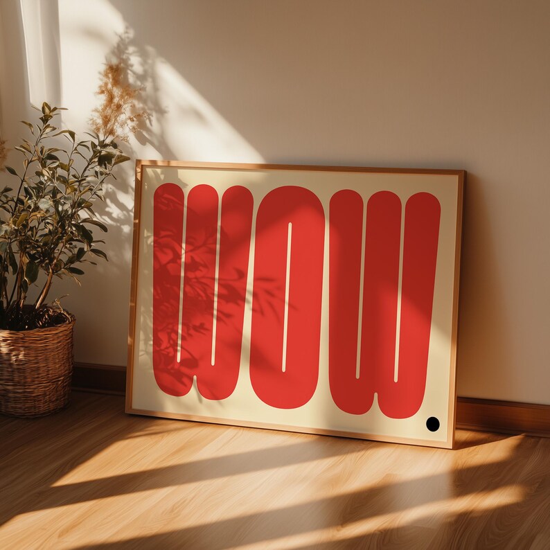 May include: A framed art print featuring the word "WOW" in bold, red, rounded letters against a cream background. The artwork is displayed on a wooden floor, leaning against a beige wall. A small black dot is in the lower right corner.