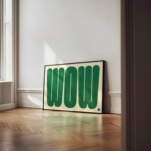 May include: A framed art print featuring the word "WOW" in bold, green, rounded letters against a cream background. A small blue dot is in the lower right corner. The artwork is displayed on a wooden floor.