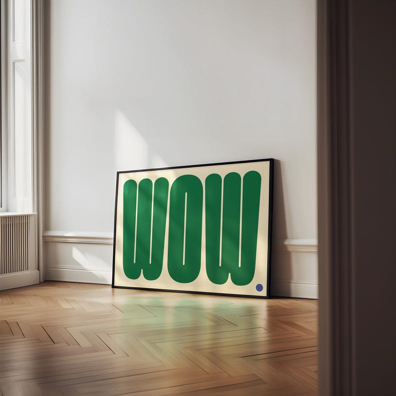 May include: A framed art print featuring the word "WOW" in bold, green, rounded letters against a cream background. A small blue dot is in the lower right corner. The artwork is displayed on a wooden floor.