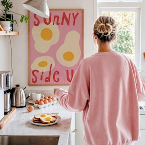 Peut inclure: Une affiche rose avec les mots "Sunny Side Up" et des illustrations d'œufs au plat est accrochée à un mur blanc. Une personne portant un sweat-shirt rose se tient dans une cuisine avec une assiette de toasts et d'œufs.