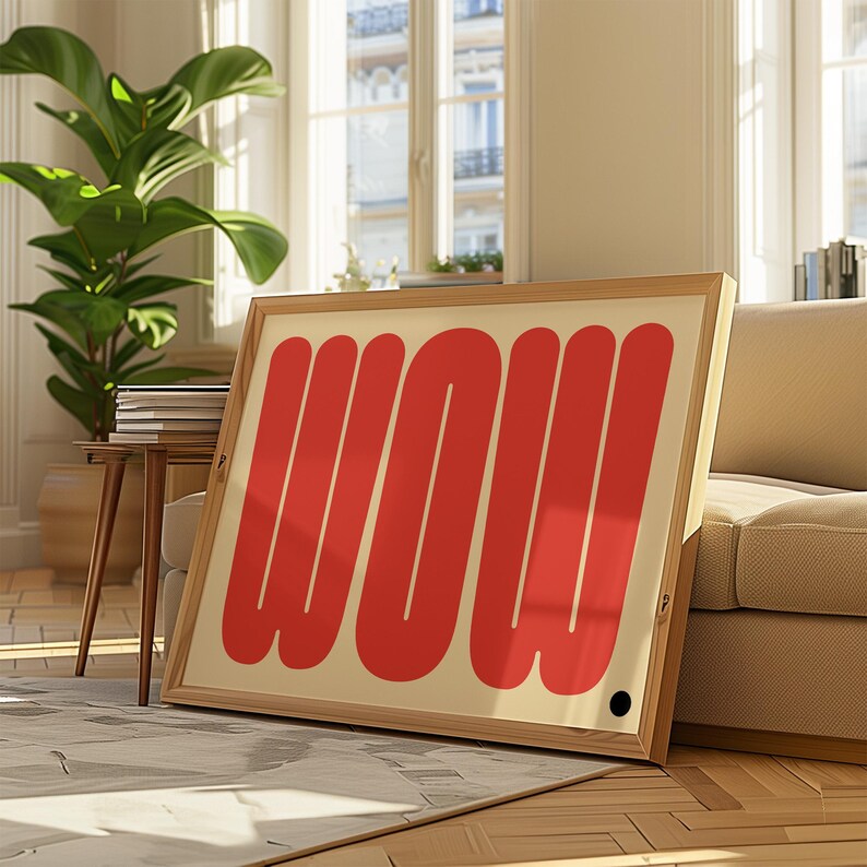 May include: A framed art print with the word "WOW" in bold, red letters. The artwork is set against a cream background and encased in a light wood frame. The print is displayed in a well-lit room with a sofa and a potted plant.