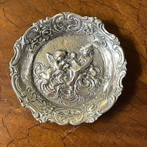 Puede incluir: Un plato de plata para alhajas con un borde adornado y festoneado. El centro presenta un diseño en relieve de dos querubines tocando una lira.