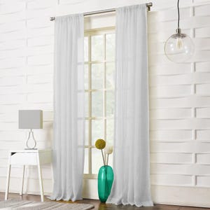 May include: White sheer curtains frame a window, set against a textured white wall. A grey lamp rests on a white bedside table. A clear glass globe pendant light hangs from the ceiling. A teal vase holds decorative flowers.
