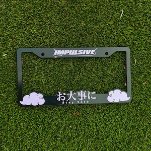 May include: A black license plate frame with the word "IMPULSIVE" in white script at the top. The bottom of the frame has Japanese text and the words "Stay Safe" in white. Two white clouds with purple outlines are on the bottom.