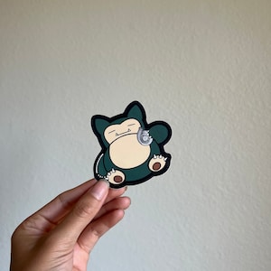 May include: A sticker featuring a cartoon Snorlax character in a relaxed pose. The Snorlax is primarily dark green, cream, and brown, with a silver stethoscope. The sticker is held by a hand against a plain background.