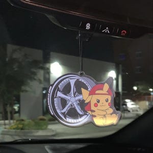 May include: A car air freshener featuring a cartoon Pikachu wearing a red cap, next to a silver car wheel design. The air freshener is hanging from a black string.