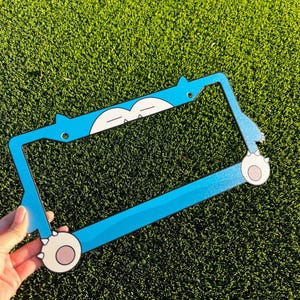 May include: A light blue license plate frame with a cartoon character design. The frame is rectangular with rounded edges and a glittery finish. The design includes white and light brown accents.