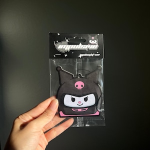 May include: A black air freshener in a clear plastic package. The air freshener is shaped like a cartoon character with a pink skull and a white face. The package has the word "impulsive" in white text.