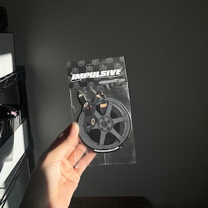 May include: A black and gray car wheel-shaped sticker featuring a cartoon character with black fur, red eyes, and tan accents. The sticker is in a clear plastic package with the word "IMPULSIVE" printed on the top.