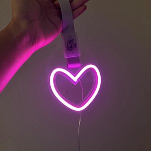 May include: A neon pink heart-shaped light with a white outline. The light is suspended by a gray strap with a small cartoon bunny logo. The light is plugged into a wall.