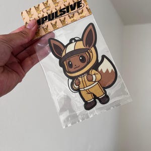 May include: A clear plastic package containing a cartoon Eevee character wearing a yellow flight suit and helmet. The packaging has a repeating pattern of Eevee characters and the word "IMPULSIVE" in black.