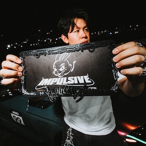 May include: A black rectangular license plate frame featuring a raised, metallic-style barbed wire design along the border. The center displays a white graphic of a rabbit head above the word IMPULSIVE in a bold, stylized white font.