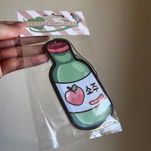 May include: A green, red, and pink air freshener in the shape of a soju bottle. The air freshener has a peach graphic and the word "소주" in Korean. The air freshener is in a clear plastic package with a pink and white striped header that says "Impulsive".
