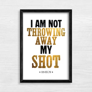 Puede incluir: Impresión enmarcada con el texto "I AM NOT THROWING AWAY MY SHOT" en letras negras y doradas. La palabra "HAMILTON" está debajo. La impresión está en un marco negro y sobre un fondo de madera blanca.
