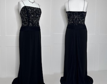 May include: A black evening gown with a beaded bodice and spaghetti straps. The bodice is embellished with silver-toned floral designs. The skirt is floor-length and flows into a small train. The dress is displayed on a mannequin.
