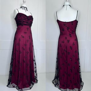 Op de afbeelding: Een vloerlange jurk met een diep magenta satijnen basislaag bedekt met zwarte mesh-stof. De jurk heeft een hartvormige halslijn, dunne spaghettibandjes en een lijfje met geplooide mesh-details. Zwart bloemenborduursel bedekt de rok van de jurk.