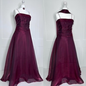 May include: A floor-length, deep burgundy gown featuring thin spaghetti straps and a fitted bodice adorned with small, scattered rhinestones. The dress has a straight neckline and a flowing, sheer-layered skirt that extends to the floor.