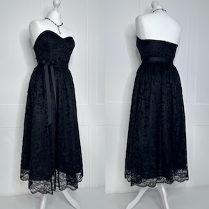 May include: A strapless black midi dress featuring an all-over floral lace overlay, a fitted bodice, and a defined waistline accented with a black ribbon tie. The skirt has a flared silhouette with a scalloped lace hemline, displayed on a mannequin.