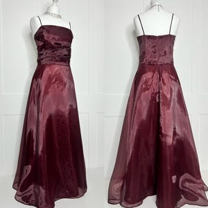 May include: A floor-length burgundy gown with a shiny, semi-sheer overlay. The bodice features thin spaghetti straps, a straight neckline, and horizontal ruched detailing with small, scattered embellishments. The skirt is full and flared.