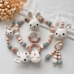 Puede incluir: Un conjunto de juguetes para bebés hechos a mano con temática de conejito. El conjunto incluye un clip para chupete, un mordedor y un sonajero, todos elaborados con ganchillo blanco, cuentas de madera y detalles en verde claro. Cada artículo está personalizado con el nombre "Thies".