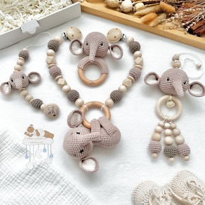 May include: A selection of baby accessories, including a pacifier clip, teether, and wooden bead garland. Each item features a crocheted elephant design in beige and brown. The accessories are made of wood and crochet.