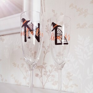 May include: Two champagne flutes with personalised Halloween-themed designs. The flutes are decorated with the letters "N" and "L" with a witch's hat and spider web design. The names "Natalie" and "Lewis" are written in orange on the glasses.