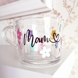 May include: A clear glass mug with a floral design and the word "Mom" in black lettering. The flowers are in shades of pink, purple, yellow, blue, and white.