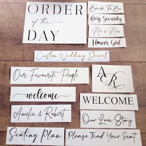 May include: A collection of white vinyl decals with black and gold lettering featuring wedding-themed phrases including ORDER OF THE DAY, Bride To Be, Ring Security, Mr & Mrs, Flower Girl, Our Favourite People, welcome, Amelia & Robert, Seating Plan, and more.