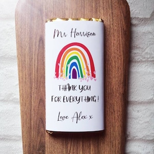 Op de afbeelding: Een chocoladereep met een witte wikkel met een regenboogontwerp en de tekst "Mr Harrison, Thank You For Everything! Love Alex x." De reep ligt op een houten snijplank met een decoratief lint.